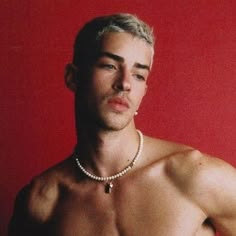 a shirtless man with no shirt on posing in front of a red wall wearing a pearl necklace