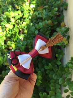 a hand holding up a red and white bow tie with scissors on the end, in front of some bushes