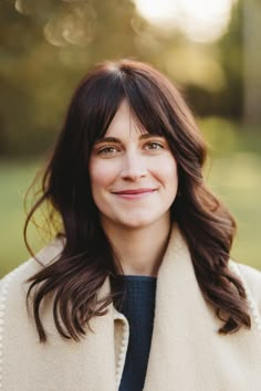 a woman with long dark hair wearing a white coat and smiling at the camera while standing in a park