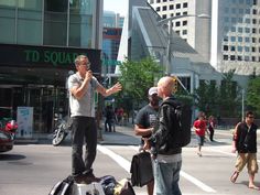 three men standing on the side of a road talking to each other while holding luggage