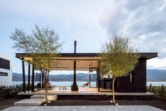 a modern house with an outdoor dining area next to the water and trees in front of it