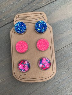 three pairs of pink and blue glittered earrings on top of a cardboard box with wooden background