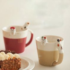 two mugs and a piece of cake on a white plate with cats figurines