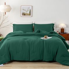 a bed with green sheets and pillows in a white room next to a lamp on a table