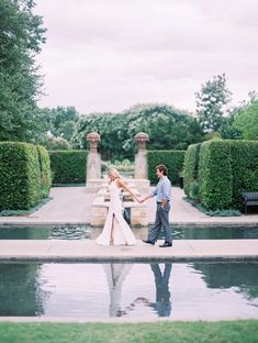 a bride and groom holding hands in front of a fountain surrounded by greenery on a cloudy day