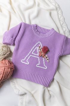 a purple sweater with the letter a on it next to two skeins of yarn