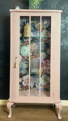 a pink china cabinet with glass doors and floral wallpaper on the walls behind it