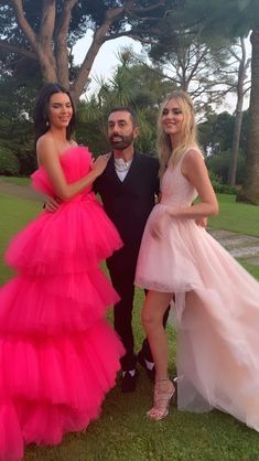 two women and a man standing next to each other in front of trees wearing pink dresses