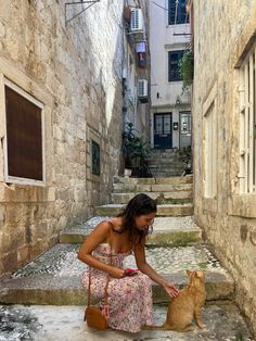 a woman kneeling down petting a cat in an alleyway with stone steps leading up to the door