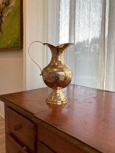 a brass vase sitting on top of a wooden table next to a window with curtains