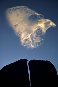 two large rocks in front of a blue sky with white clouds above them and one rock sticking out from the ground