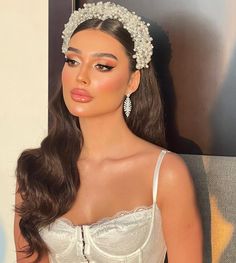 a woman with long brown hair wearing a white dress and headpiece, standing in front of a wall