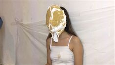 a woman wearing a white tank top standing in front of a wall with a gold mask on it's head