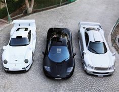 three white sports cars parked next to each other on a cobblestone road with trees in the background