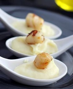 three white spoons filled with food on top of a black plate and topped with scallops