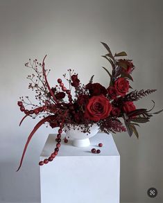 a vase filled with red flowers and greenery on top of a white pedestal next to a wall