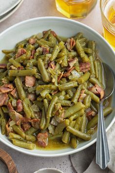 a bowl filled with green beans and bacon next to two silver spoons on a table