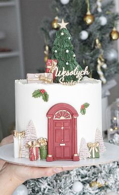 a white christmas cake with red door and presents on the top, sitting in front of a decorated christmas tree