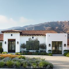 a white house with lots of plants and shrubs in front of the building that is surrounded by mountains