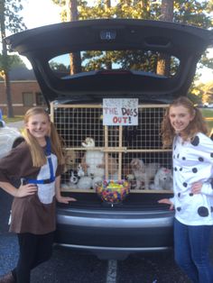 two girls standing next to the back of a car with dogs in cages on it