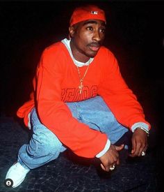 a young man sitting on the ground wearing an orange sweatshirt and blue jeans with white sneakers