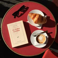 two plates with croissants and a book on a red round table outdoors