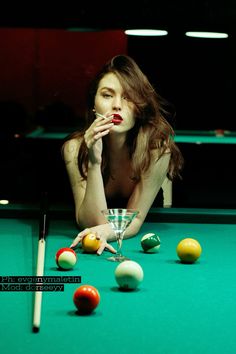 a woman leaning over a pool table with a martini in her hand and billiard balls on the floor