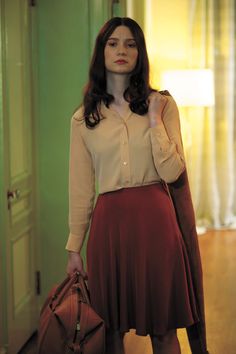 a woman standing in a hallway holding a handbag and looking off into the distance