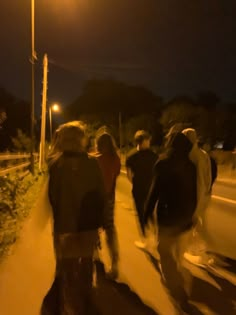 a group of people walking down a sidewalk at night time with street lights in the background