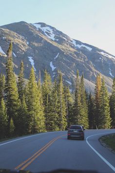 a car is driving down the road in front of some trees and snow covered mountains