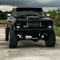 a large black truck parked on top of a cement parking lot next to tall grass