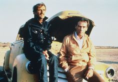 two men sitting on top of an old car in the desert, one is holding his leg up