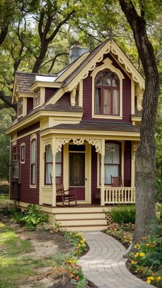 a small yellow and red house in the middle of some trees with flowers around it