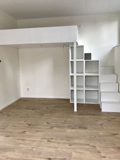 an empty room with white shelves and wooden floors in the center is a ladder leading up to stairs