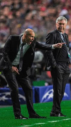 two men in suits standing on a soccer field with one pointing at the other man