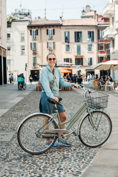 a woman standing next to a bike on a cobblestone street with buildings in the background