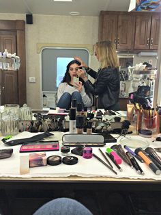 two women sitting at a table with makeup and other items on it in front of a mirror