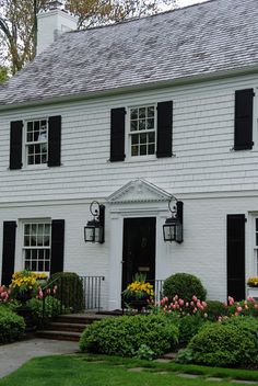 a large white house with black shutters and flowers in the front yard on a cloudy day