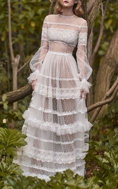 a woman standing in the woods wearing a white dress with long sleeves and tiered layers