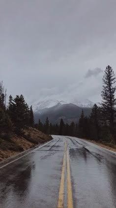 an empty road with snow capped mountains in the distance and trees on both sides, under a cloudy sky