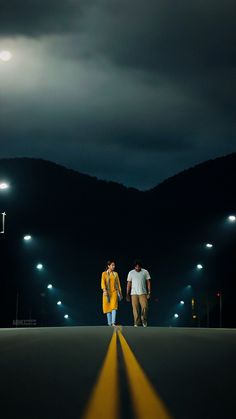 two people are walking down the road at night with lights shining on mountains in the background