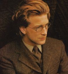 a man wearing glasses and a suit looks off into the distance with his hand on his chest