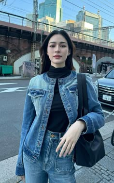 a woman standing on the side of a road wearing a jean jacket and black turtle neck top