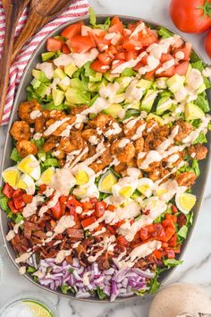 a large salad with dressing in a bowl on a marble countertop next to tomatoes, lettuce, cucumbers and other vegetables