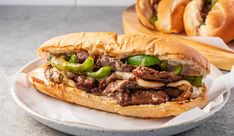 a beef sandwich with peppers and onions on a white plate next to bread buns