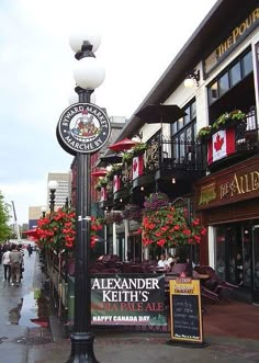 a lamp post with flowers on it in front of a building and people walking down the street