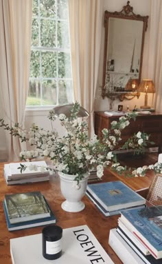 a table topped with books and vase filled with white flowers next to a large window