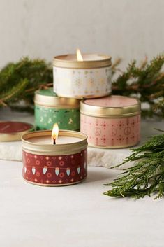 four candles sitting next to each other on top of a white table with pine branches