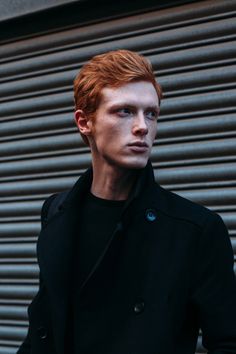 a man with red hair wearing a black coat and standing in front of a building