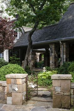 a stone fence and gate in front of a house with trees, shrubs and bushes